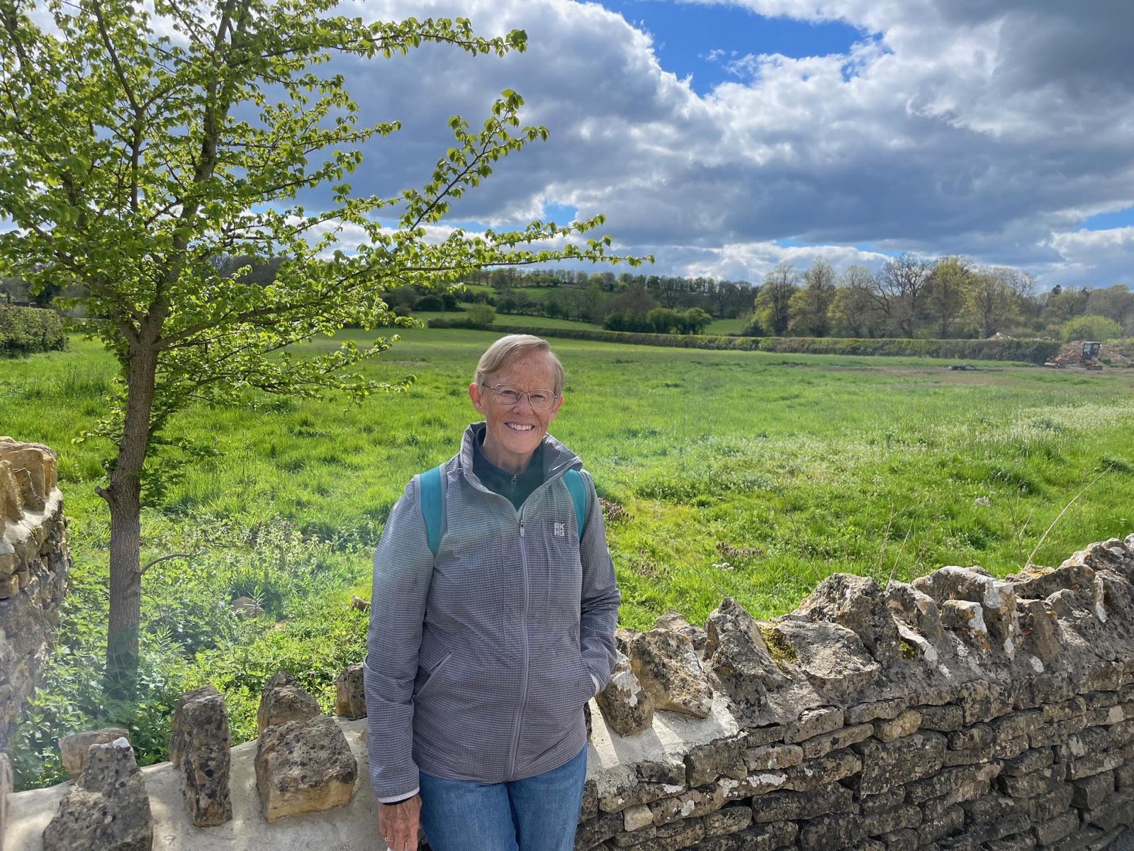 Mary Jean smiling beside a dry stone wall with rolling green Cotswolds fields behind her